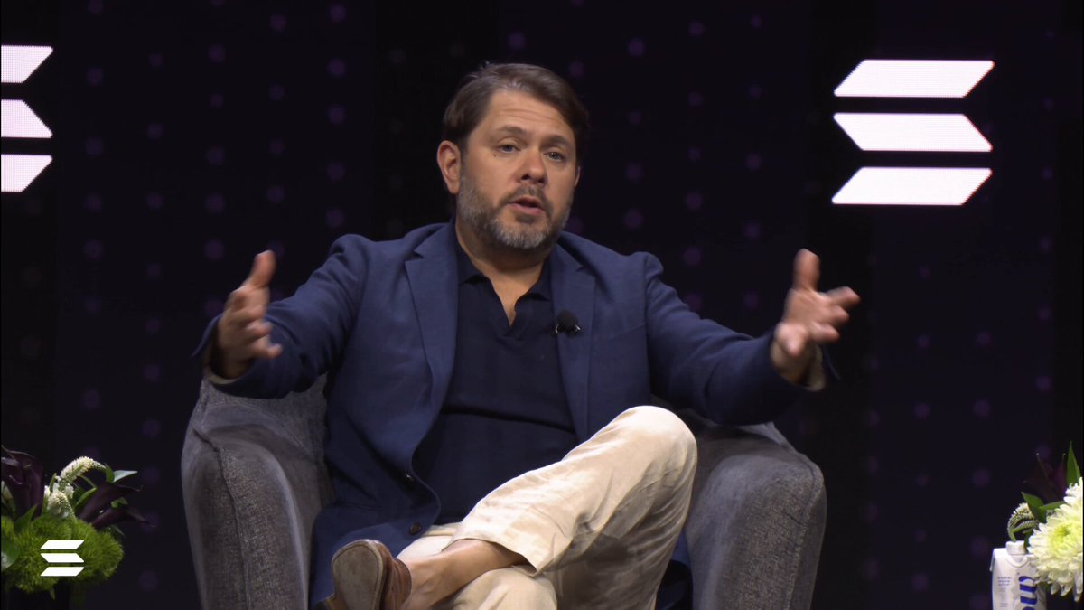The image features a man sitting in a chair, wearing a blue suit jacket and white pants. The background of the image shows a purple wall with white letters that say "S", which could be interpreted as an advertisement or promotional material for Sberbank. The caption provided below the image reads "Why does the US need Clarity? Here’s Sen. @RubenGallego". This suggests that the man in the suit jacket is speaking at a conference, possibly addressing the importance of clarity and transparency in the financial industry.

The image also includes a potted plant placed on one side, which adds some greenery to the scene. The presence of the plant might indicate an effort to create a more pleasant atmosphere for the event or presentation where the man is speaking.