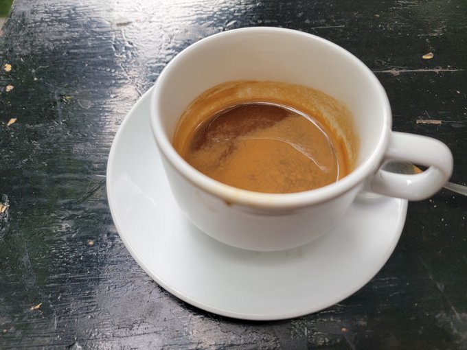 A small white ceramic espresso cup and saucer sit on a weathered, dark wooden table, with the cup containing the dregs of a finished coffee.