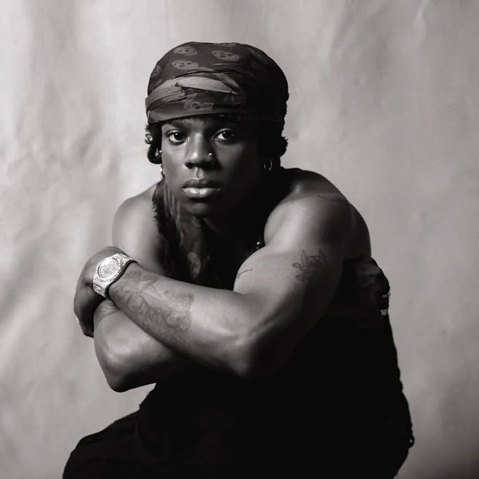 A black and white portrait of a person wearing a bandana with circular patterns, displaying visible tattoos on their arms while wearing a watch.