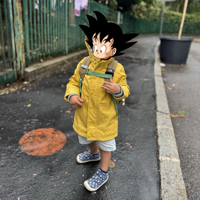 A child wearing a yellow raincoat and backpack stands on a paved path, with their head replaced by an animated character featuring spiky black hair resembling Goku from Dragon Ball.