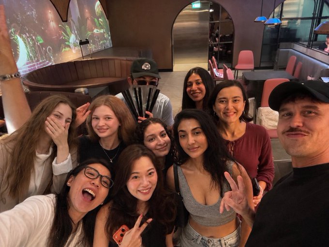 A group selfie of nine people posing together in what appears to be a modern lounge or office space, with one person holding up chopsticks.