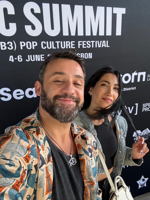 A man and woman take a selfie in front of a backdrop reading "C SUMMIT" and "(B3) POP CULTURE FESTIVAL," with the man wearing a colorful patterned shirt and the woman in a denim jacket, capturing the celebratory moment at the Bitcoin art event where they are building BTCArtSociety together.