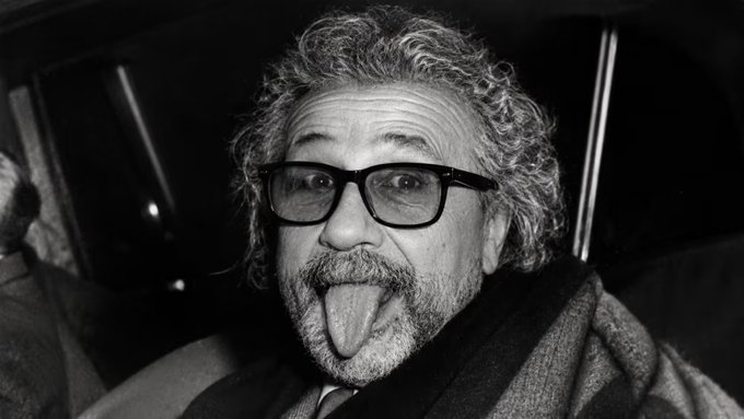 A black-and-white close-up shows a curly-haired, bearded man wearing glasses and sticking out his tongue playfully at the camera. The quirky, exaggerated expression captures the playful "meme" spirit referenced in the accompanying caption about "Supeinstein," a humorous mashup meme created by @tinoch and Jedi.