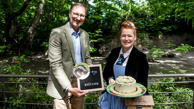 A man in a tan checkered blazer and a woman in a blue checkered dress stand outdoors in a wooded setting, holding a letter board reading 'BRÖTCHEN' with hearts and a plate displaying a green frosted cake.