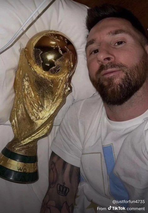 Lionel Messi lies in bed next to the FIFA World Cup trophy, smiling contentedly in a white t-shirt with visible arm tattoos. The TikTok watermark "@justforfun4735" and "From TikTok comment" tag appear in the corner, accompanying a post by the user celebrating with the caption "Happy Akara Tuesday Mortals."