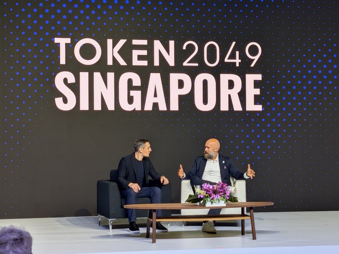Two men seated in armchairs on a stage engaged in conversation at the TOKEN 2049 SINGAPORE conference, with a coffee table and purple flowers between them.