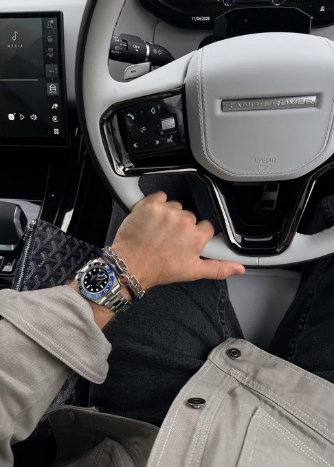 A first-person view from the driver's seat of a Range Rover, showing a hand resting on the steering wheel wearing a black and blue Rolex Submariner watch.