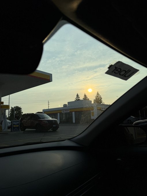 A view from inside a vehicle looking out through the windshield at a Shell gas station (with a reversed 'open' sticker on the glass) during sunrise or sunset, with a dark SUV parked at the pumps.