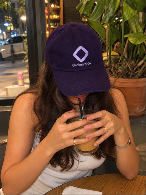 A woman wearing a navy baseball cap branded with "@nebula2004" takes a sip from a glass with a straw at what appears to be a café or restaurant, with greenery in the background. The image pairs with her announcement of being booked by @monad to cover their flagship conference during TOKEN2049 week in Singapore, showcasing her creator-ready aesthetic.