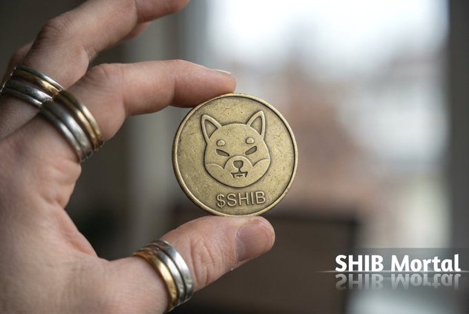 A hand adorned with silver rings holds a gold-colored coin featuring the Shiba Inu dog emblem and the text "$SHIB," symbolizing the tangible rise of memecoins from internet joke to legitimate financial asset. The watermark "SHIB Mortal" appears in the corner, reinforcing the post's message that $SHIB represents community-driven culture merging with finance.