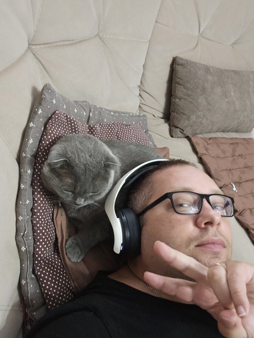 A selfie of a man wearing glasses and white headphones, lying on a tufted couch next to a grey cat resting on a brown polka-dot pillow.