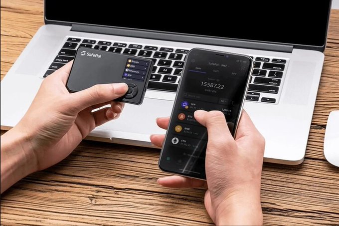 Two hands hold a SafePal hardware wallet and a smartphone displaying the SafePal crypto app—showing a balance of 1558722 CYP/USDT alongside portfolio holdings like SXP, ARDR, AVAX, and ETH—over a MacBook keyboard, illustrating a user managing digital assets on hardware and mobile devices that have been linked to SafePal's recent data breach affecting nearly 39,798 customers.
