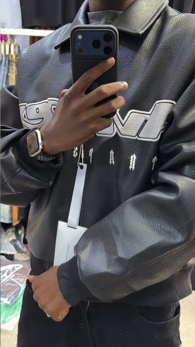 A mirror selfie of a person wearing a black leather jacket with white stylized lettering, holding an iPhone, with a smartwatch on their wrist and a white strap hanging from their neck.