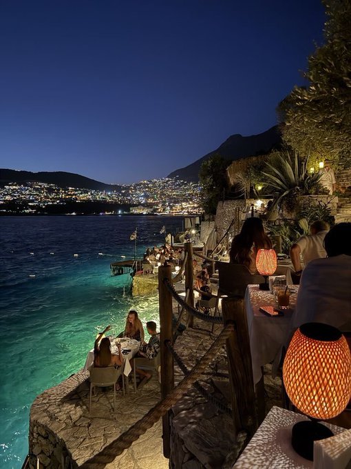 A coastal restaurant terrace glows with warm orange lanterns against the deep blue night sky, where diners enjoy seaside tables overlooking a lit-up bay and hillside city.

The serene, intimate evening scene reflects the post's theme of winding down and preparing for rest, evoking the kind of peaceful nightcap moment that precedes a meaningful night's sleep.