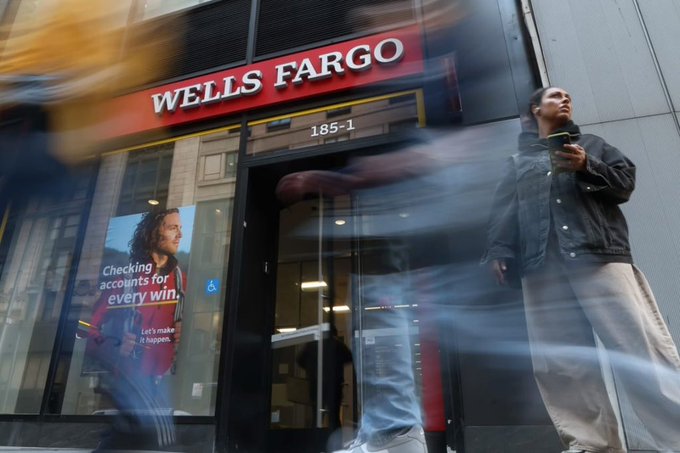 A Wells Fargo bank branch at address 185-1 displays an advertisement reading "Checking accounts for every win" and "Let's make it happen," while blurred pedestrians and a man holding a coffee cup walk past the storefront, visually representing the financial institution at the center of the news about its new blockchain-based tokenized deposit initiative.