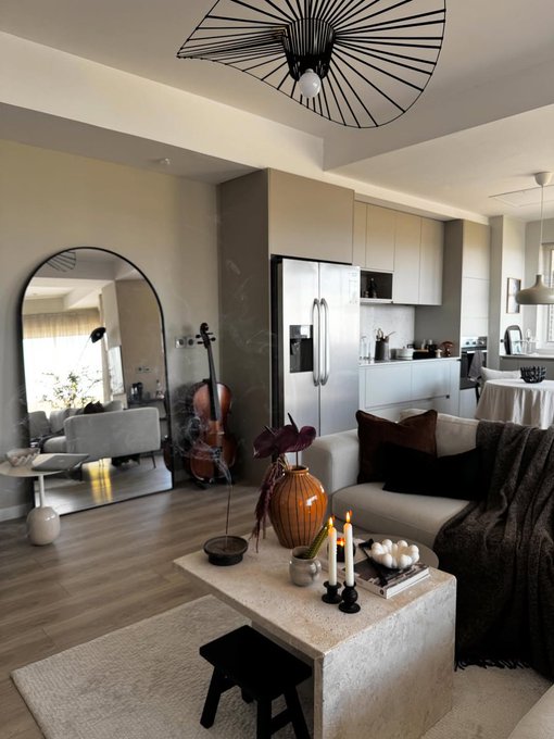 A stylish open-plan living room features a neutral-toned sofa with dark accents, a stone coffee table styled with candles and decorative objects, and a dramatic arched mirror reflecting a sunlit seating area. A striking geometric chandelier hangs from the ceiling, while a cello and kitchen area are visible in the background, creating a warm, artistic atmosphere.
