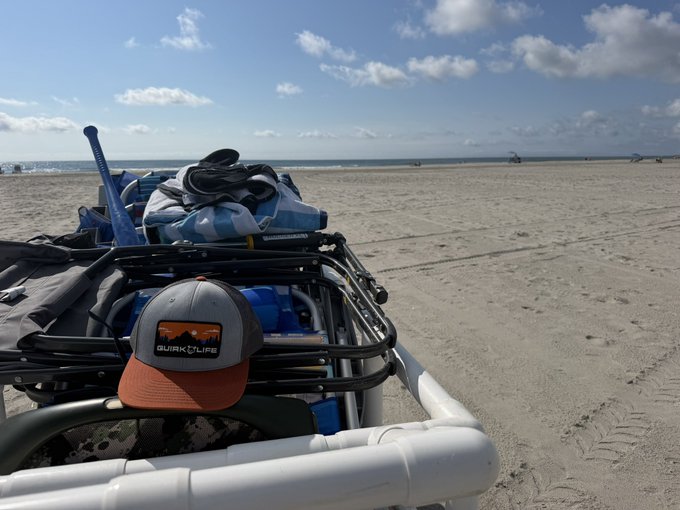A first-person view from a beach cart loaded with gear, featuring a brown and gray 'QUIRK LIFE' hat in the foreground, looking out over the sandy shore toward the ocean under a partly cloudy sky.