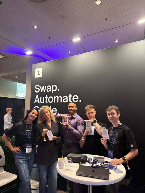 The image shows a group of five people standing around a table, posing for a photo with their laptops open. They are all wearing name tags that say "swap.automeate". The backdrop behind them features a black wall with white text that reads "swap.automeate", and there is also a sign that says "1" on it. A laptop can be seen on the table, along with some cups placed nearby.