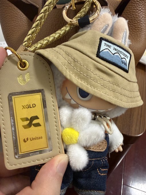 A plush doll wearing a bucket hat with 'THE MONSTERS' patch and denim overalls, attached to a keychain with an 'XGLD Unitag' tag.