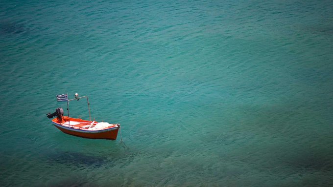 An aerial view of a small red and white boat with a Greek flag floating in clear turquoise water.
