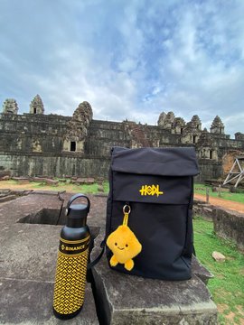 A travel photo showing a black backpack with a yellow star keychain and a yellow and black water bottle placed on stone, with ancient temple ruins (likely Angkor Wat/Bayon) in the background under a blue sky.