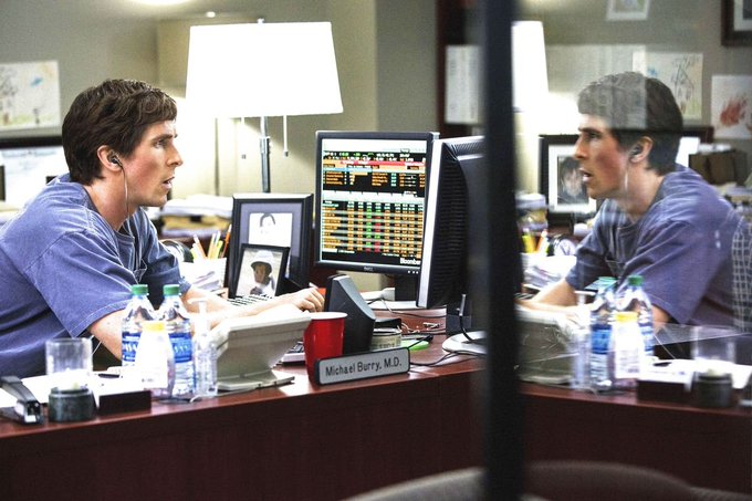 A man in blue scrubs works at a desk with multiple computer monitors displaying medical data, with a nameplate reading 'Michael Berry, M.D.' and his reflection visible in a glass surface.