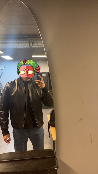The image features a man wearing a black leather jacket and sunglasses taking a selfie with his cell phone, which has a green filter applied to it. The background of the photo includes a mirror that reflects the man's face along with some furniture such as a chair and a desk. There is also an object in the room that appears to be a suitcase or a box.