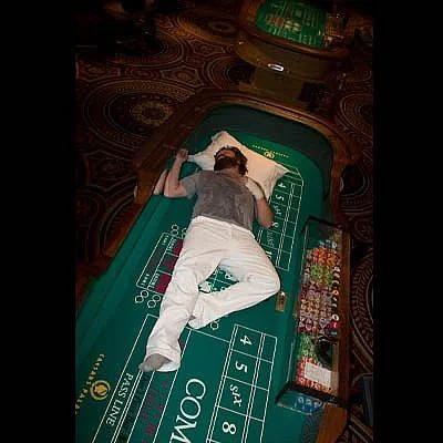 A person sleeping on a casino craps table at what appears to be a Las Vegas venue, wearing white pants and resting their head on a pillow placed directly on the gaming felt.