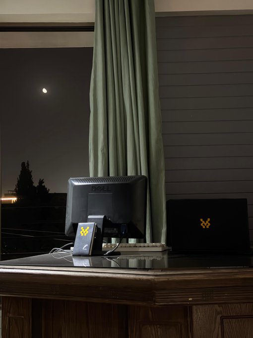 A desk setup at night with a monitor, a laptop with an illuminated orange logo, a small device with a yellow logo, and the moon visible through a window.