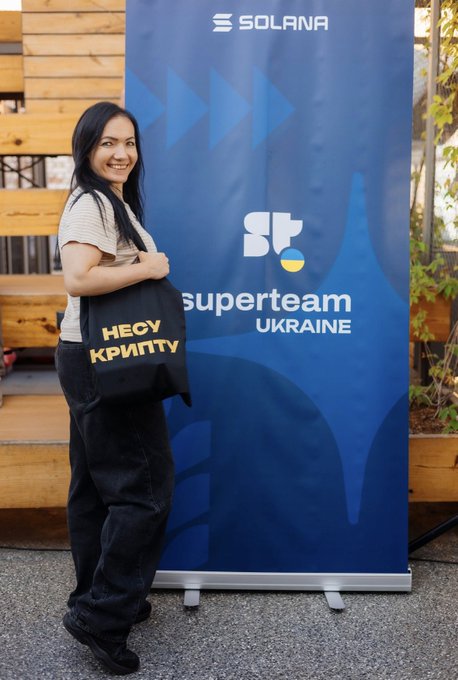 The image features a woman standing next to a blue banner that displays the text "Superteam Ukraine" and "Kyiv", possibly indicating an event or gathering related to Ukraine. The woman is holding a black bag with gold lettering, which could be a promotional item or a gift for the occasion.