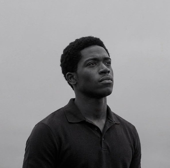 A black-and-white portrait of a young man in a dark polo shirt looking off to the side against a plain light background.