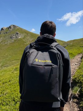 The image shows a man wearing a backpack with "binance" written on it, walking up a hill or mountain trail. He appears to be enjoying his outdoor adventure while carrying his backpack filled with items for his journey. The surrounding environment consists of grassy areas and mountains in the background, creating a picturesque setting for the man's hike.