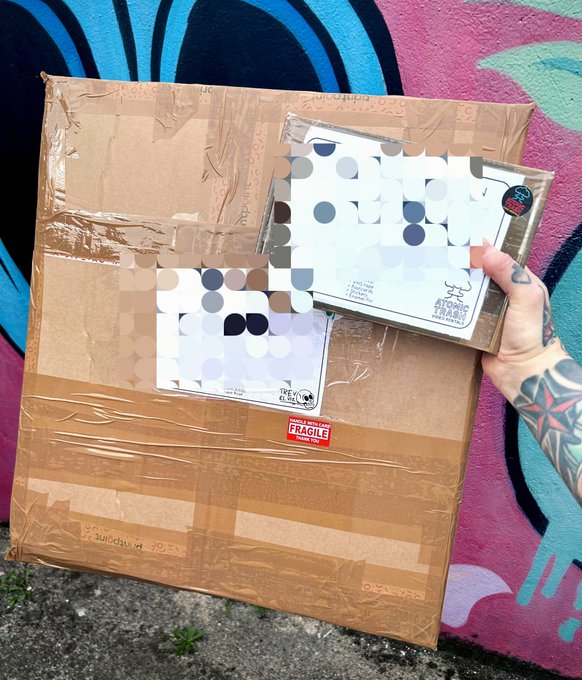 A hand with tattoos holding two Trend-Closet clothing packages over a cardboard shipping box marked 'FRAGILE' against a colorful graffiti-painted wall.