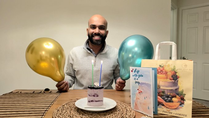 A smiling bald man with a beard sits at a table celebrating a birthday with a small cake featuring two lit candles, flanked by gold and teal balloons, with a "Life with you is a trip" greeting card beside him.
