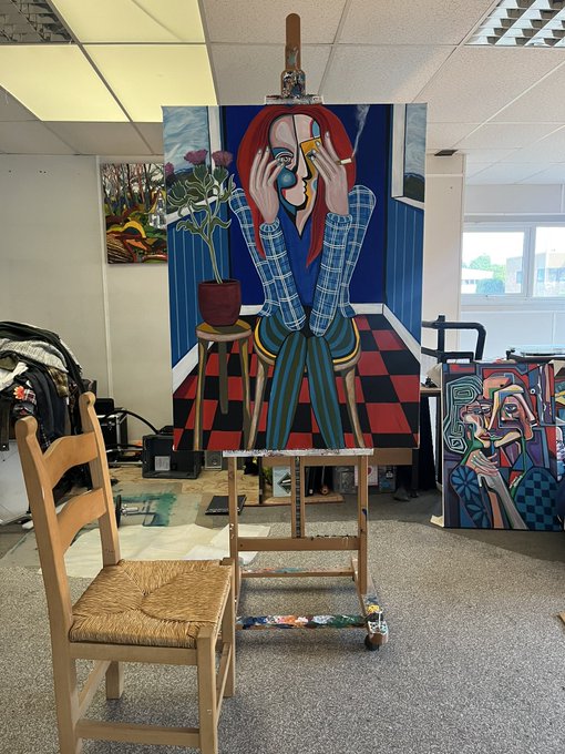 An artist's studio featuring a large cubist-style painting of a figure with red hair on an easel, alongside other artworks, a wooden chair, and an easel.