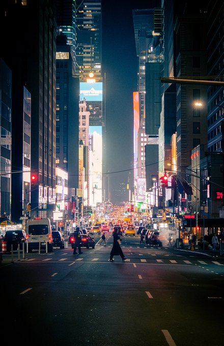A vibrant nighttime cityscape of a busy urban street, likely Tokyo, featuring tall illuminated skyscrapers, traffic lights, pedestrians crossing, and bright billboards lining the road.