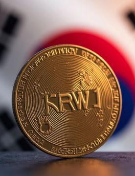 A close-up of a gold physical 'KRW1' cryptocurrency token rests on a surface in front of a blurred Korean flag.