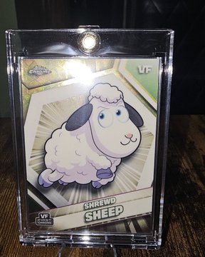 In this image, there's a clear plastic card with a cartoon sheep design sitting on top of a wooden table. The card has a silver clip holding it together at the bottom. The text "The Shrewd Sheep" is written on the front of the card in white letters. Below that, there are two lines of black text: "VF" and "Shredd Sheep". The background of the image shows a wooden table with a green surface behind the card.