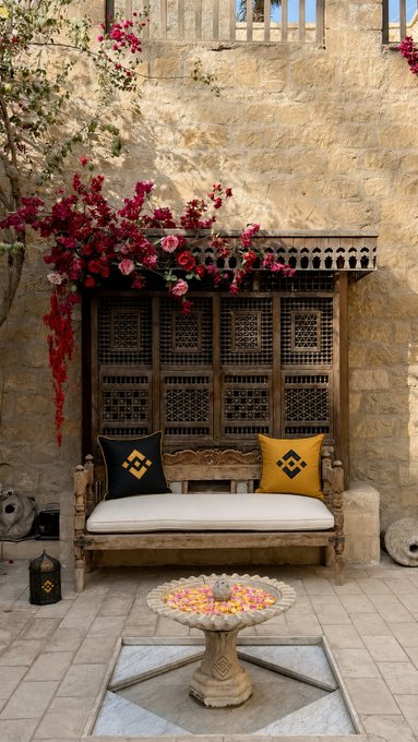 The image features an old stone building with a courtyard that has two wooden benches placed under a tree. The benches are adorned with yellow pillows, adding a touch of color to the scene. A fountain can be seen nearby, enhancing the serene atmosphere. In the background, there is a potted plant and a vase, further contributing to the overall ambiance. The image also includes text that reads "Good morning from a serene corner, its details steeped in heritage and its touch digital." This suggests that the scene might be part of an online advertisement or social media post promoting a digital platform like Binance.