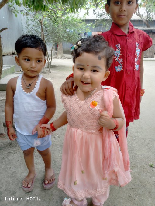 Three young children, with the smallest in the center wearing a pink dress and bindi, stand outdoors smiling while holding a lollipop, seemingly unrelated to the post's Web3 discussion of @sleepagotchi and $SLEEP tokens. The image contains a watermark reading "Infinix HOT 12" in the bottom corner, indicating the device used to capture the photo.