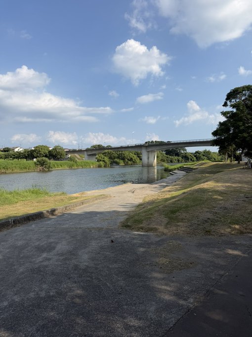 A scenic daytime view of a calm river crossed by a long concrete bridge, with a concrete boat ramp in the foreground under a bright blue sky with scattered clouds.