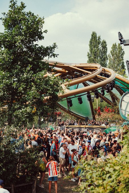 A crowd of festival-goers dances with raised hands beneath a wooden canopy stage and lush trees, embodying the post's invitation to "Let yourself be guided by the music and the @kucoincom guardians" in an outdoor music event setting.