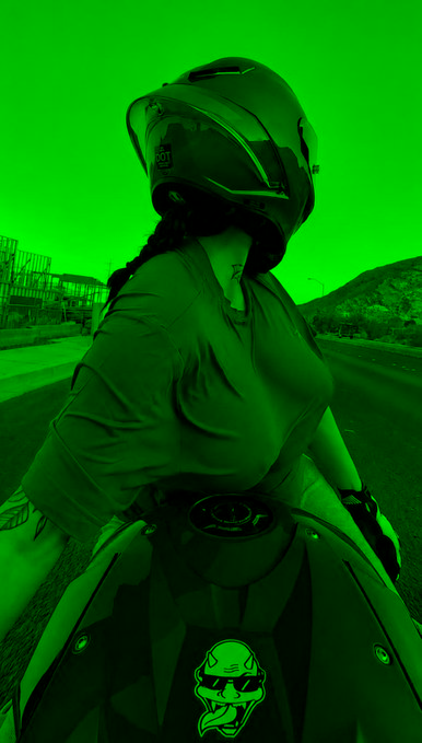 A motorcyclist wearing a full-face helmet sits on a sportbike along an open road, captured in a heavy green-tinted filter with a skull-and-tongue logo visible on the bike's fairing, fitting the post's playful suggestion that the rider—rather than the poster—is the one who belongs on the motorcycle.