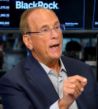 A man in glasses and a suit gestures while speaking, with a BlackRock display visible behind him. The image accompanies news of BlackRock CEO Larry Fink expressing bullish sentiment on Bitcoin, crypto, and broader markets.
