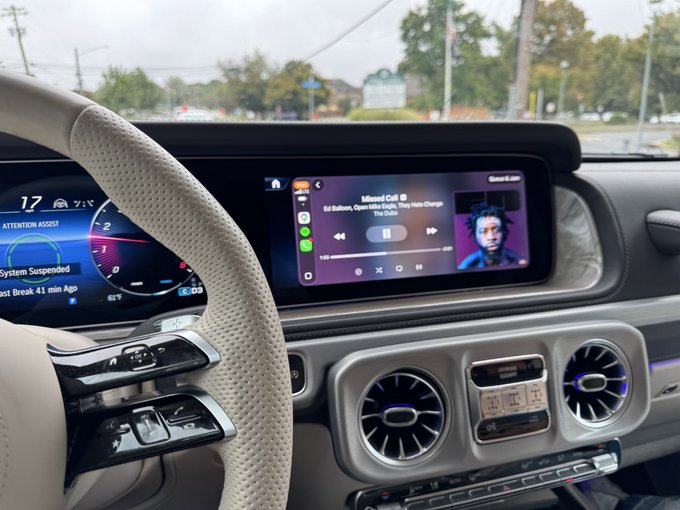 The interior view from a driver's seat shows a Mercedes dashboard displaying 'Missed Call' from Ed Ballou on the infotainment screen alongside an animated avatar, with the instrument cluster indicating 'System Suspended'.