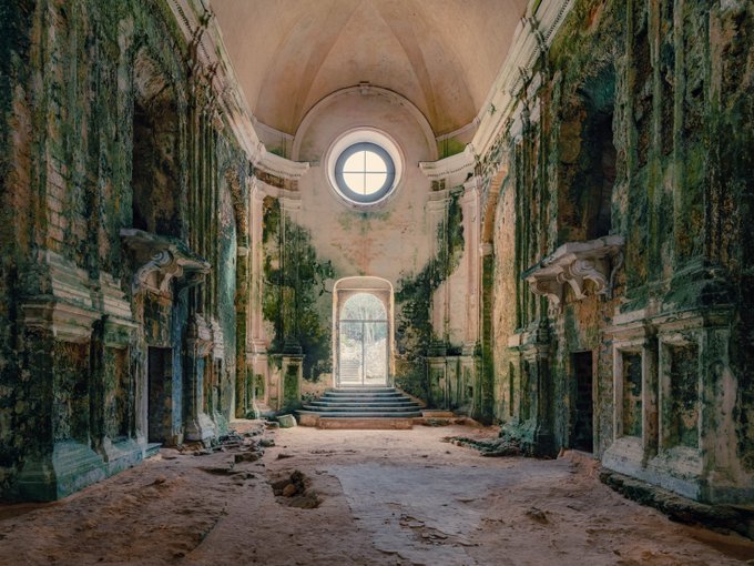 The interior of an abandoned, decaying church or building with peeling green paint, ornate stonework, a round window, and a bright doorway at the end of the hall.