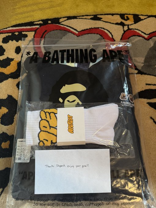 An A Bathing Ape merchandise package containing a black t-shirt with the brand's signature ape head logo, a pair of white socks with orange graphics, and a handwritten note that reads 'Thanks Sheperd, enjoy your grail!'