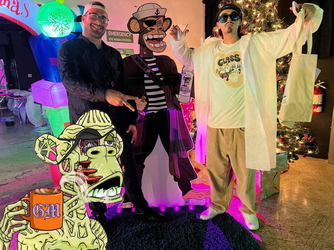 Two men pose indoors at what appears to be a Christmas event, standing next to a pirate monkey cutout and a cartoon BAPE figure, with a decorated tree in the background.