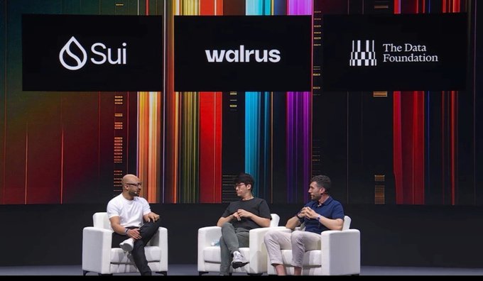 Three men sit in white armchairs on a stage in front of colorful screens displaying the logos for 'Sui', 'Walrus', and 'The Data Foundation'.