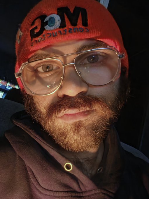 A close-up selfie of a bearded man wearing glasses and a red beanie with 'M Constuction' (mirrored) text, taken in dim lighting.